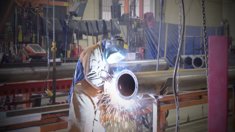 A welder joining steel pipe with sparks flying in an industrial fabrication shop